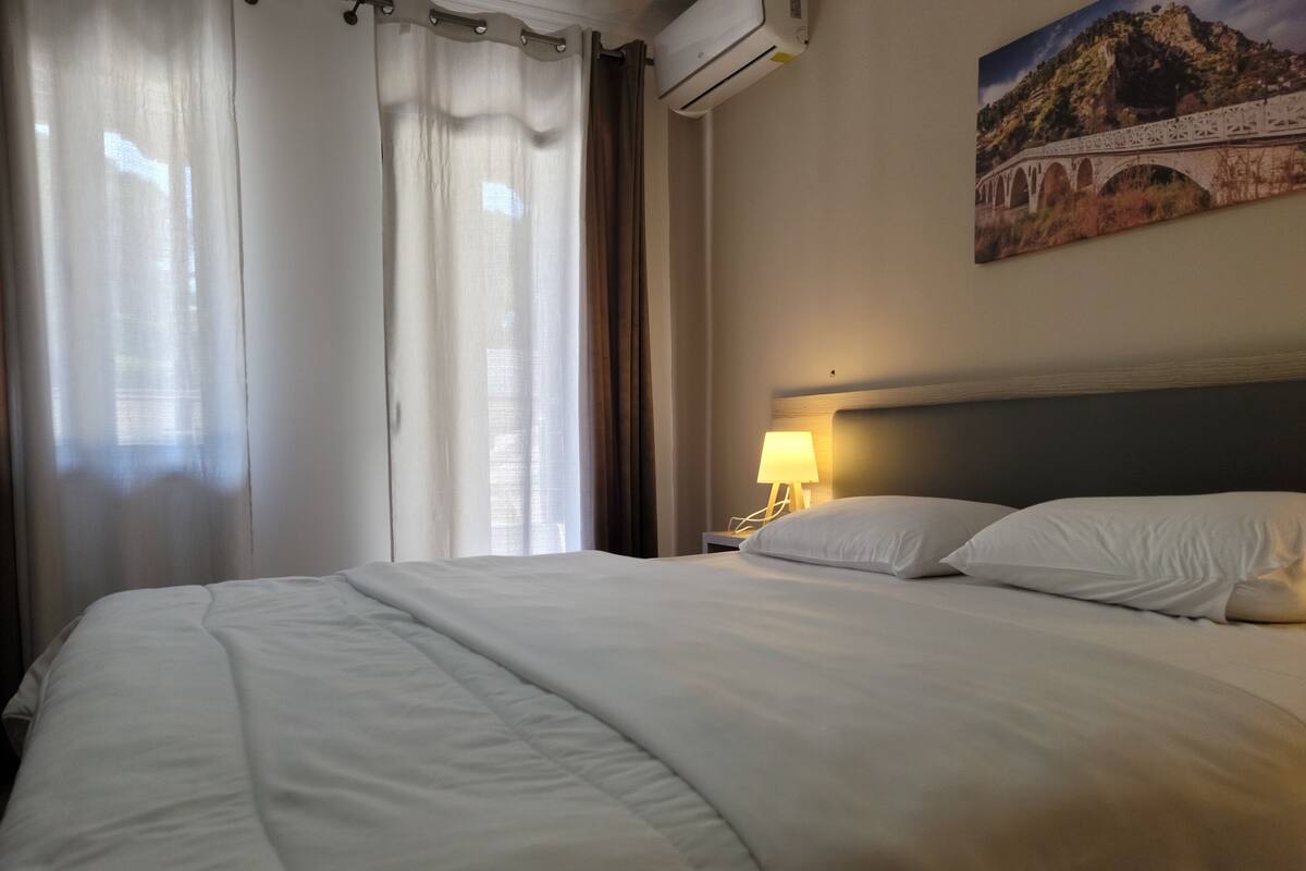 A hotel room with a neatly made bed, a nightstand with a lamp, and a painting of a bridge on the wall.