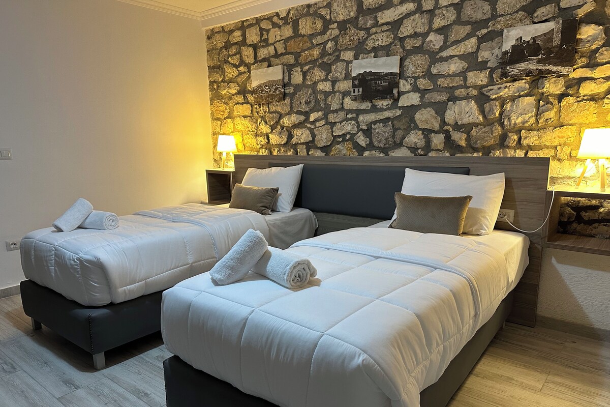 Habitación doble con dos camas, cada una con una almohada y una toalla enrollada. Hay dos mesitas de noche con lámparas. La habitación tiene una pared de piedra con cuadros enmarcados.