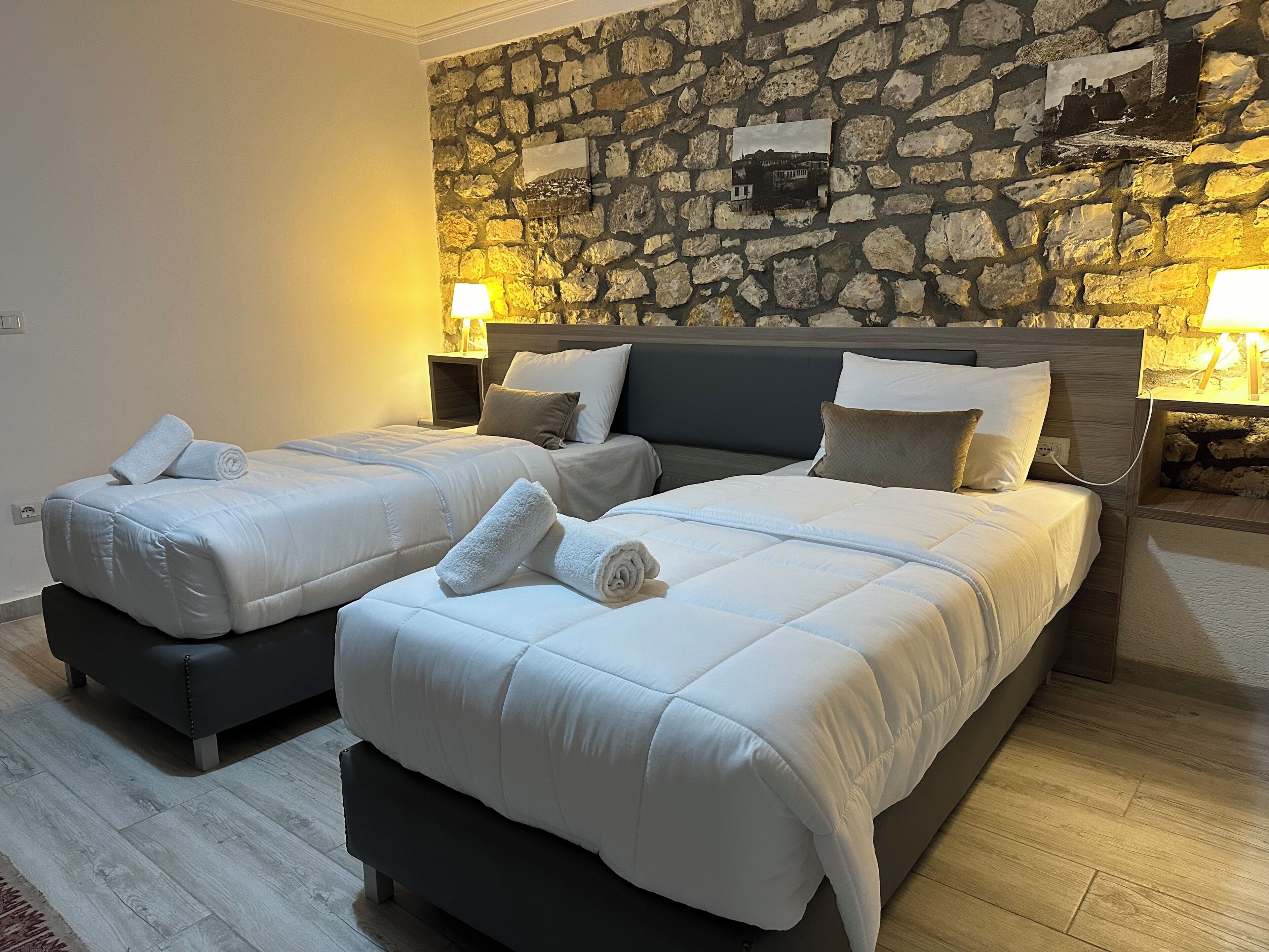 A double room with two beds, each with a pillow and a rolled towel. There are two bedside tables with lamps. The room has a stone wall with framed pictures.