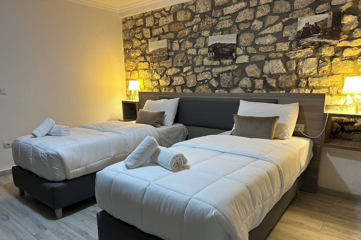 Habitación doble con dos camas, cada una con una almohada y una toalla enrollada. Hay dos mesitas de noche con lámparas. La habitación tiene una pared de piedra con cuadros enmarcados.