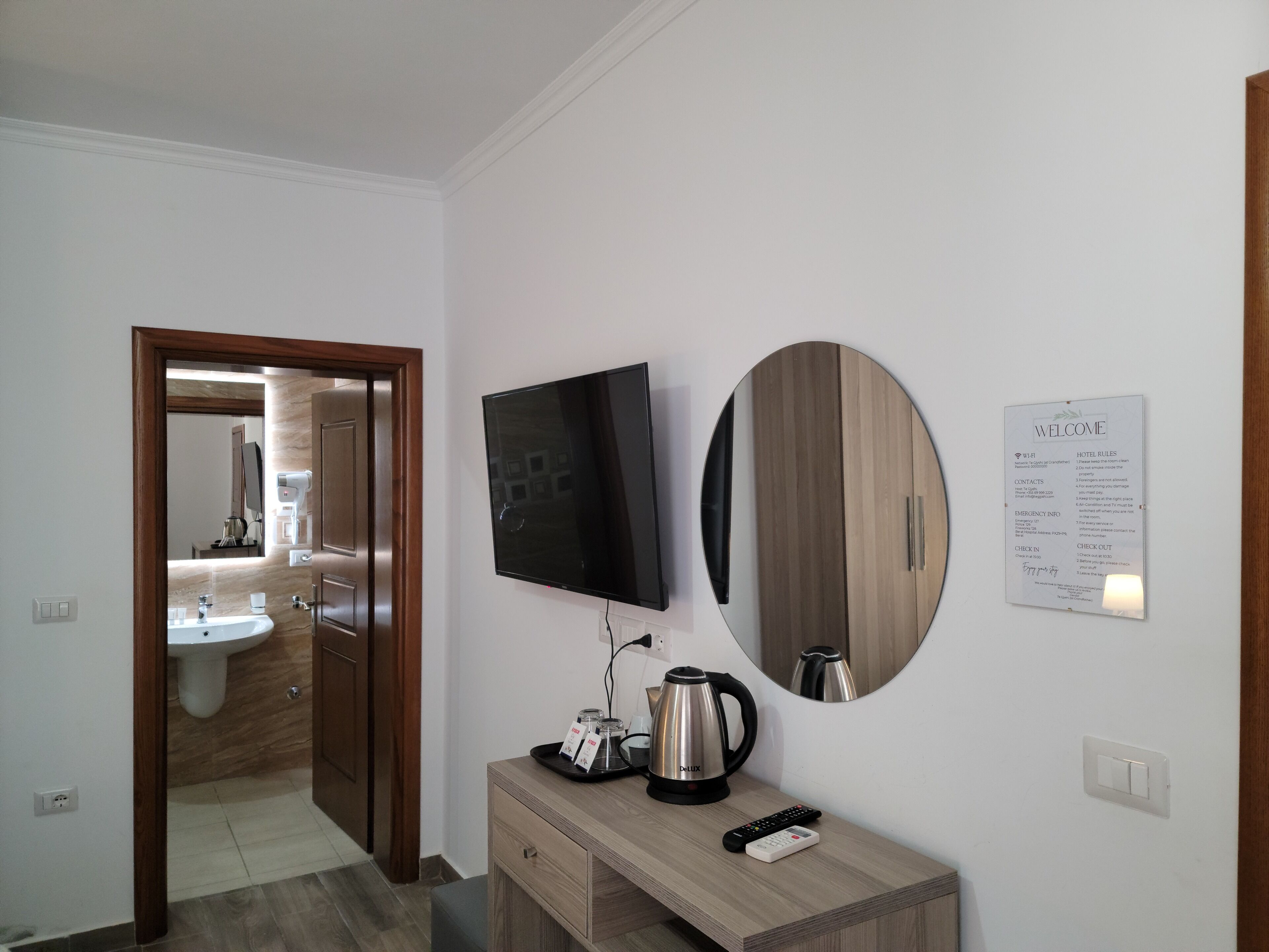 Quarto de hotel com televisão de tela plana, espelho redondo, cafeteira, controle remoto e um letreiro com regras do hotel e informações de emergência.