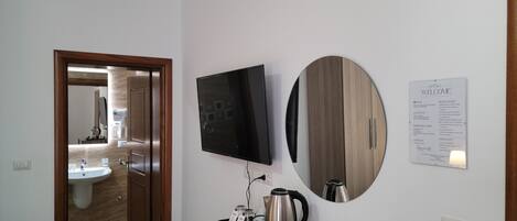 Quarto de hotel com televisão de tela plana, espelho redondo, cafeteira, controle remoto e um letreiro com regras do hotel e informações de emergência.