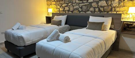Habitación doble con dos camas, cada una con una almohada y una toalla enrollada. Hay dos mesitas de noche con lámparas. La habitación tiene una pared de piedra con cuadros enmarcados.