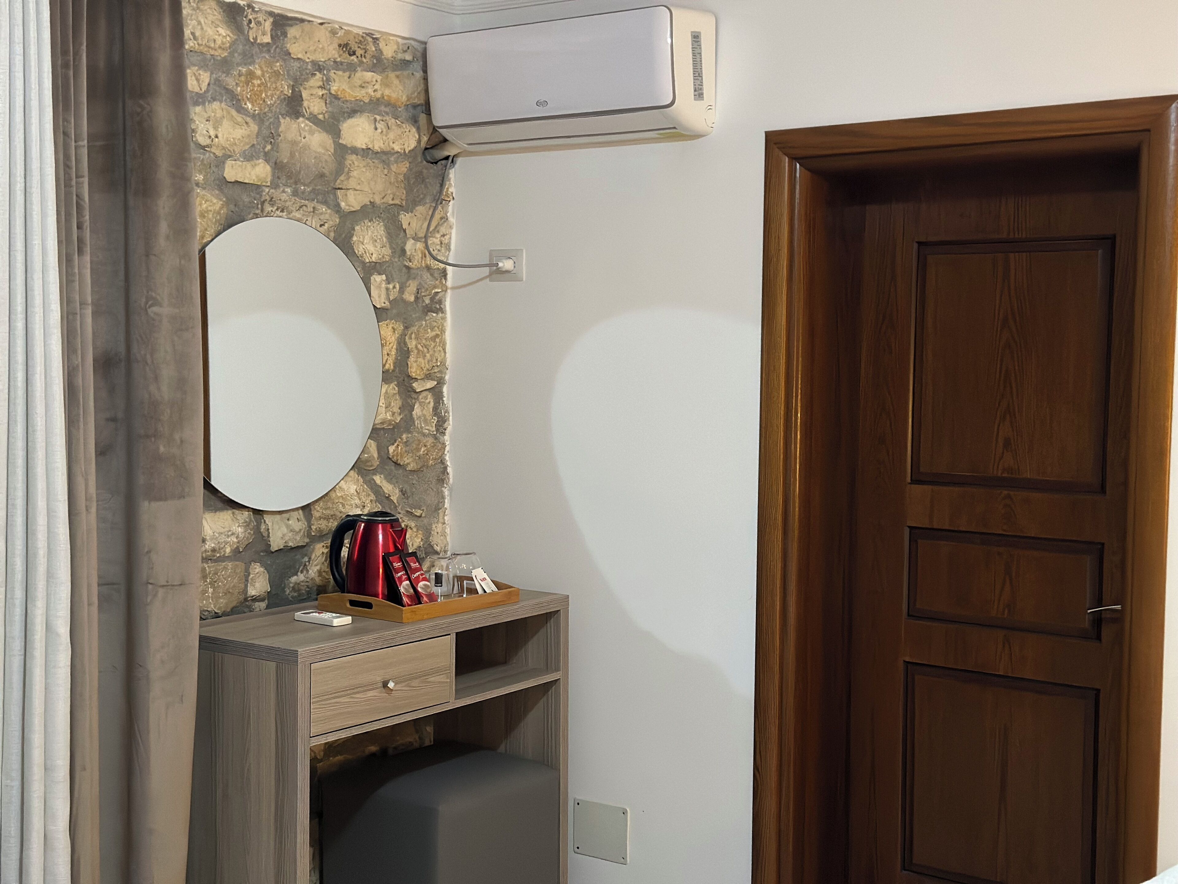 A bathroom with a stone wall, a mirror, a wooden cabinet, a red kettle, and a wooden door.