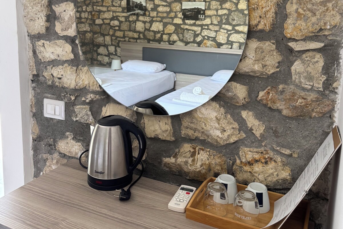Pared de piedra con un espejo redondo que refleja una cama y una tetera sobre un tocador de madera.
