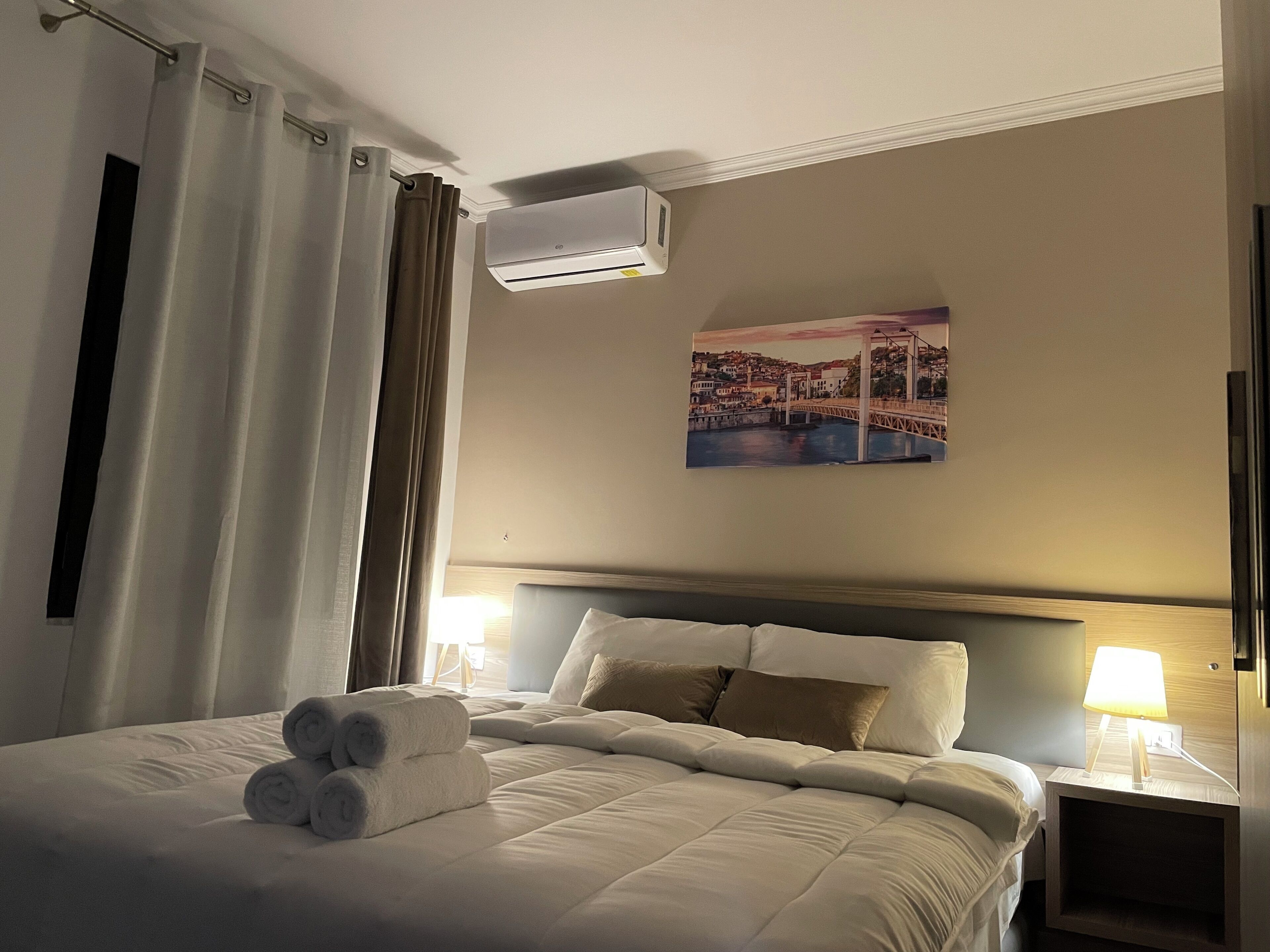 A hotel room with a bed, a headboard, a wall-mounted air conditioner, a painting of a bridge, and a nightstand with a lamp.