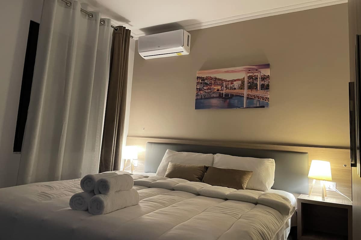A hotel room with a bed, a headboard, a wall-mounted air conditioner, a painting of a bridge, and a nightstand with a lamp.