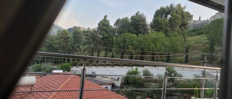 A view from a balcony with a metal railing, overlooking a red-tiled roofed area and a green landscape with trees and a hill in the background.