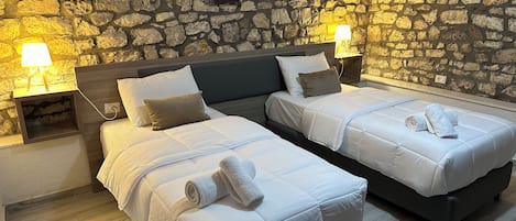 A double room with two beds, each with white bedding and a rolled towel. The room features a stone wall and two bedside tables with lamps.