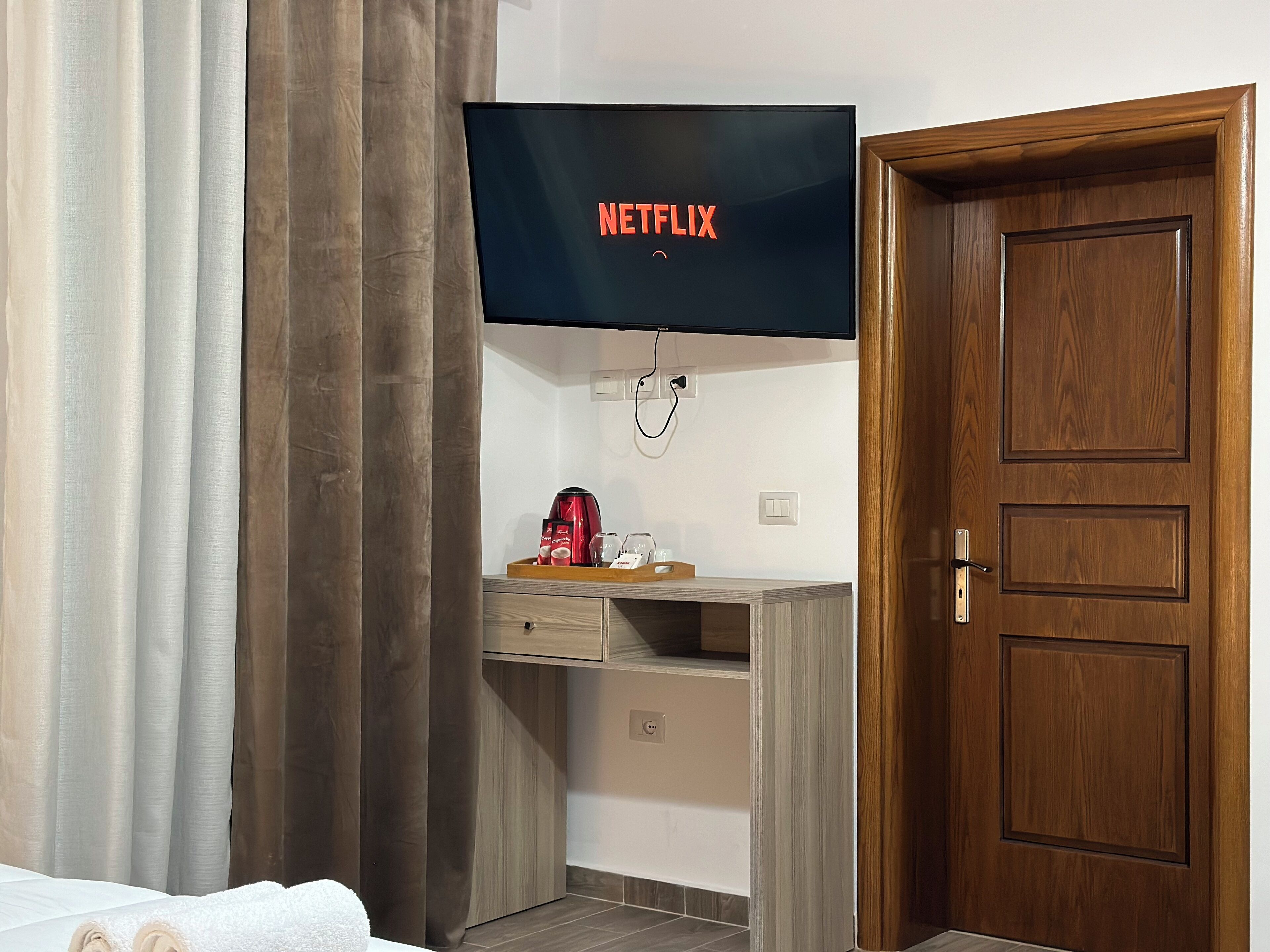 Une pièce dotée d’un téléviseur à écran plat affichant le logo de Netflix, d’un bureau en bois sur lequel se trouve un plateau avec divers objets, d’une porte en bois et d’un lit recouvert de draps blancs.