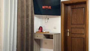 A room with a flat-screen TV displaying the Netflix logo, a wooden desk with a tray of items, a wooden door, and a bed with white linens.