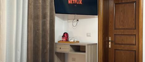 A room with a flat-screen TV displaying the Netflix logo, a wooden desk with a tray of items, a wooden door, and a bed with white linens.