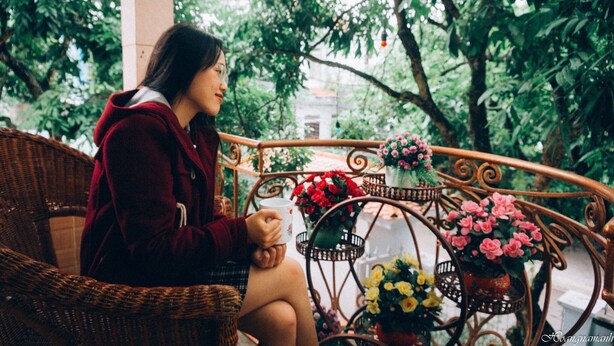 A person sitting on a wicker chair holding a cup, with a metal railing and potted flowers in the background. - Sen's homestay (Nam Hoa Lu)