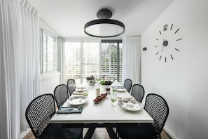 A dining room with a white table, black chairs, a large wall clock, and a window with blinds.