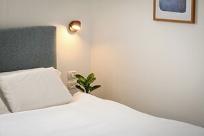 A neatly made bed with white linens, a grey headboard, a bedside lamp, a power outlet, and a small green plant.