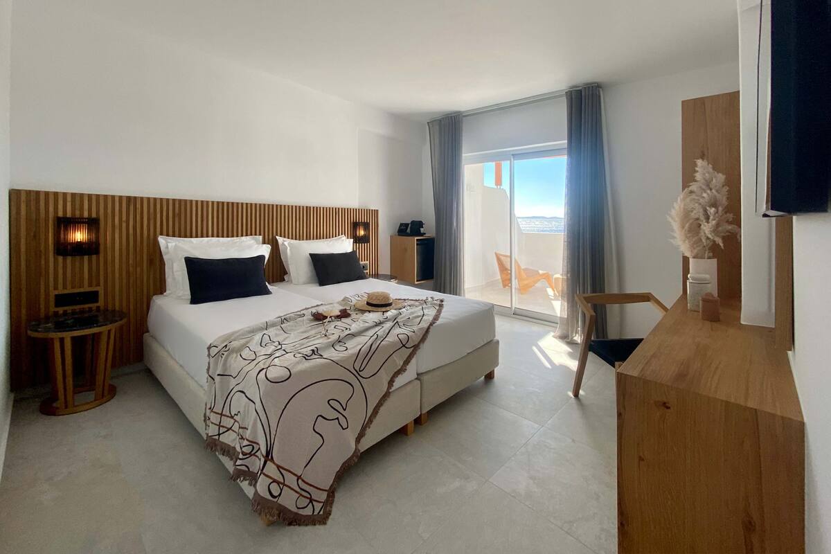 A modern hotel room with a large bed, a wooden headboard, a small table, a chair, and a view of the outside through a sliding glass door.