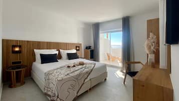 A modern hotel room with a large bed, a wooden headboard, a small table, a chair, and a view of the outside through a sliding glass door.