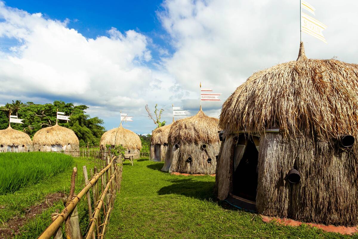 Thatched-roof huts with flags, arranged in a row with a bamboo fence in the foreground.