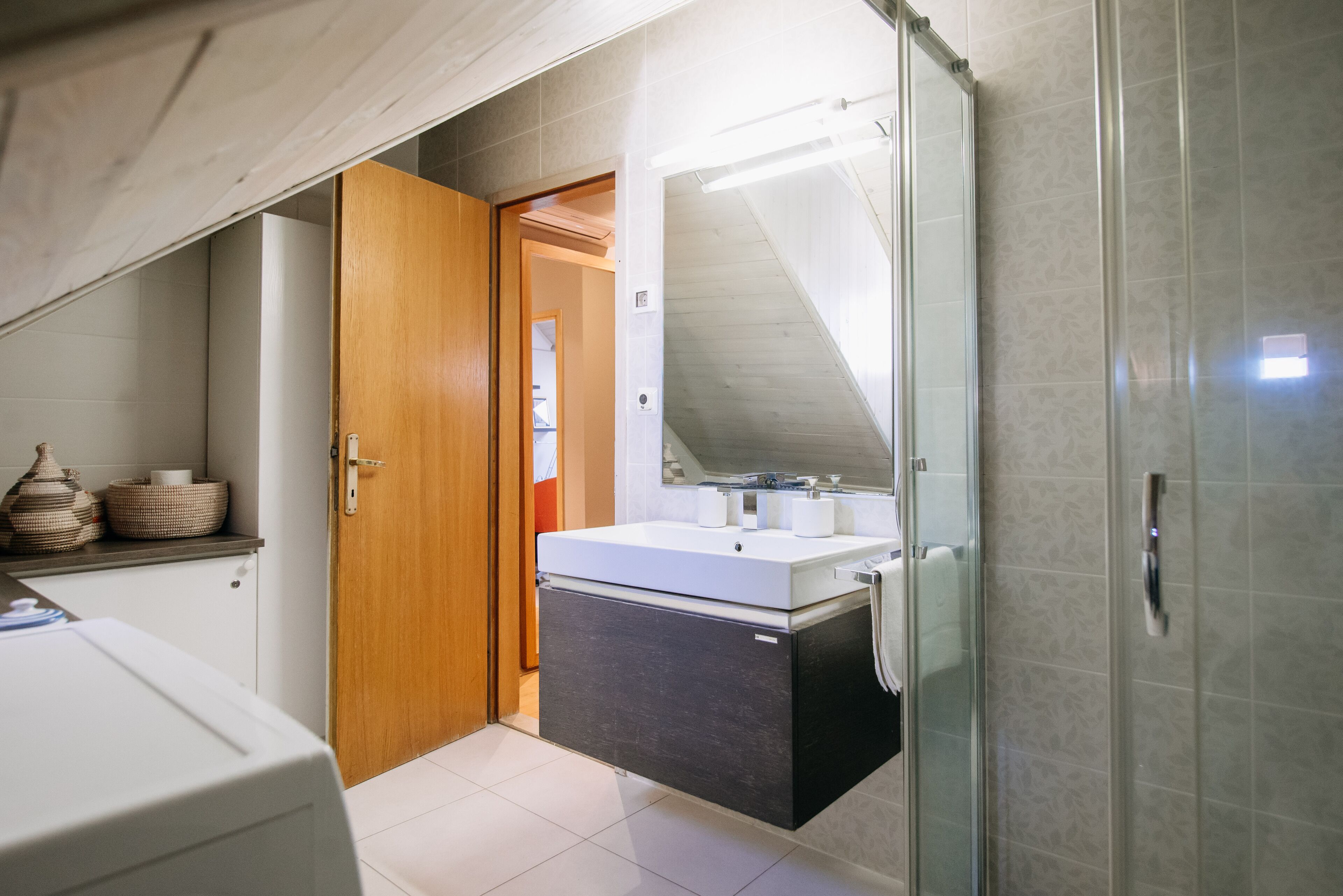 A compact bathroom with a shower, a sink with a mirror, and a wooden door leading to another room.