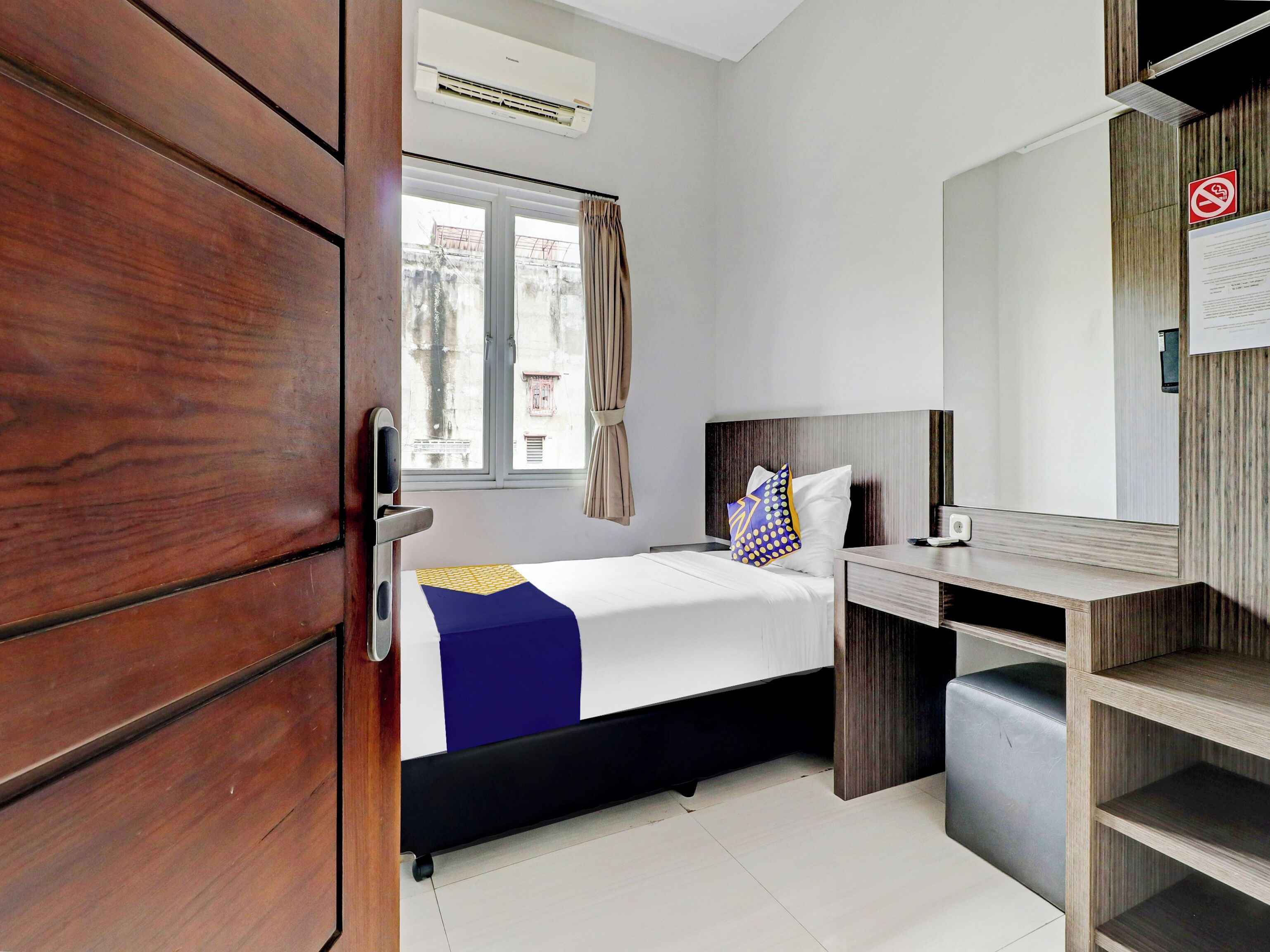 A hotel room with a bed, a desk, a chair, a wardrobe, a window with curtains, and an air conditioning unit.