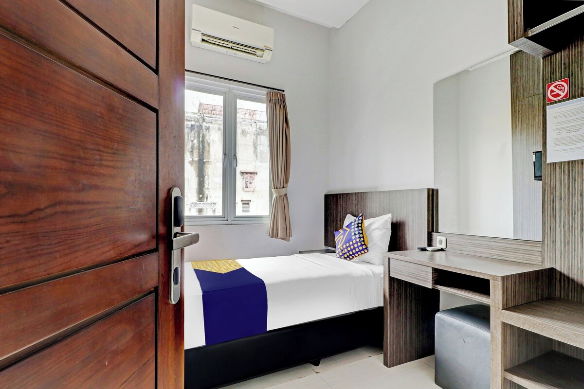 Una habitación de hotel con cama, escritorio, silla, armario, ventana con cortinas y unidad de aire acondicionado.