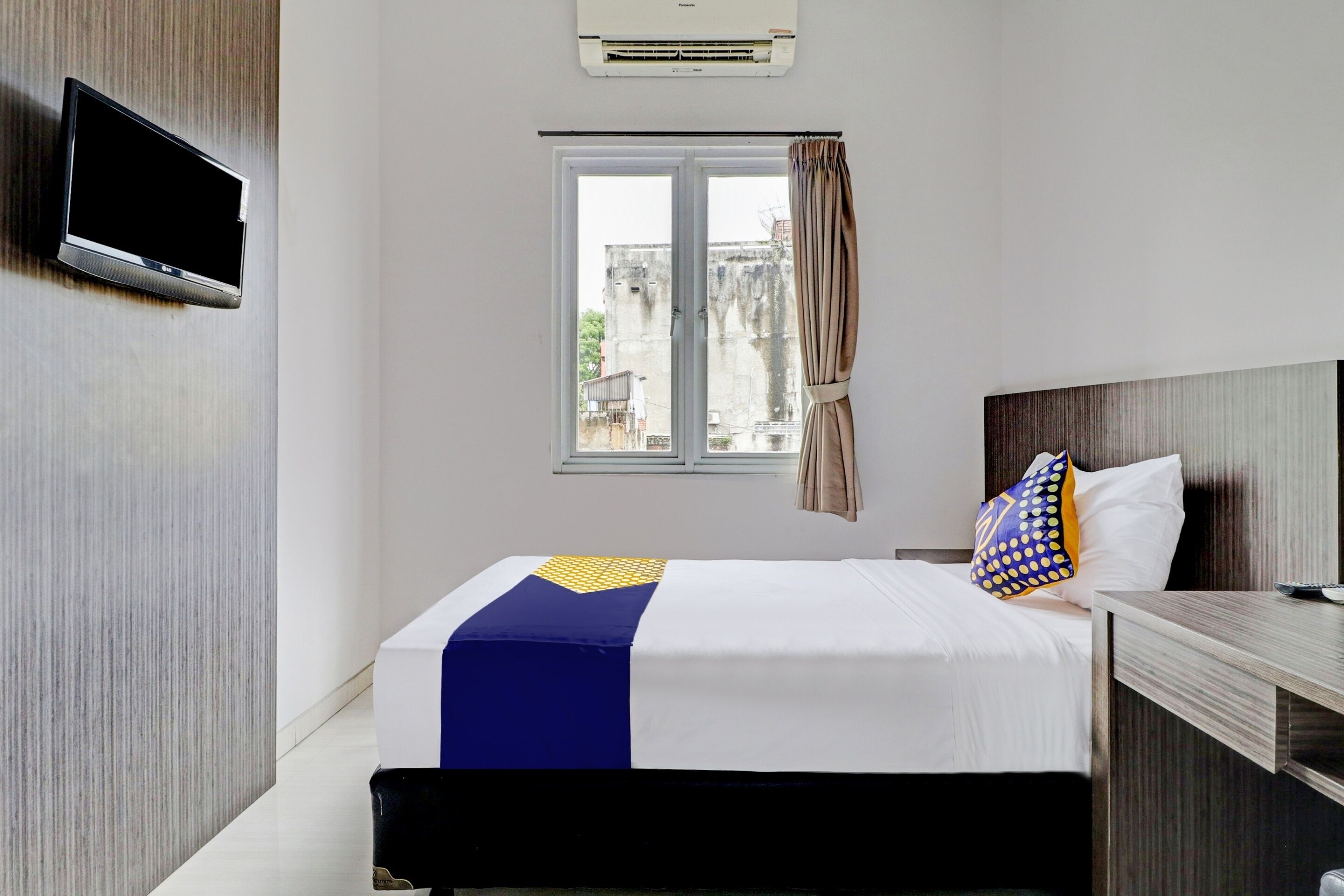 Een hotelkamer met een bed, een aan de muur bevestigde televisie, een raam met gordijnen en een uitzicht op gebouwen.
