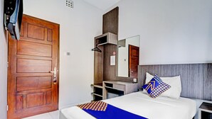 A hotel room with a wooden door, a bed with white bedding and a blue accent, a bedside table, a mirror, and a wardrobe. - Hotel O Cihampelas Near Ciwalk Formerly D'River Guest House (Bandung)