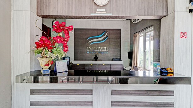 A reception desk with a clock, a logo for 'D'RIVER Guest House', and a potted plant with red flowers. - Hotel O Cihampelas Near Ciwalk Formerly D'River Guest House (Bandung)