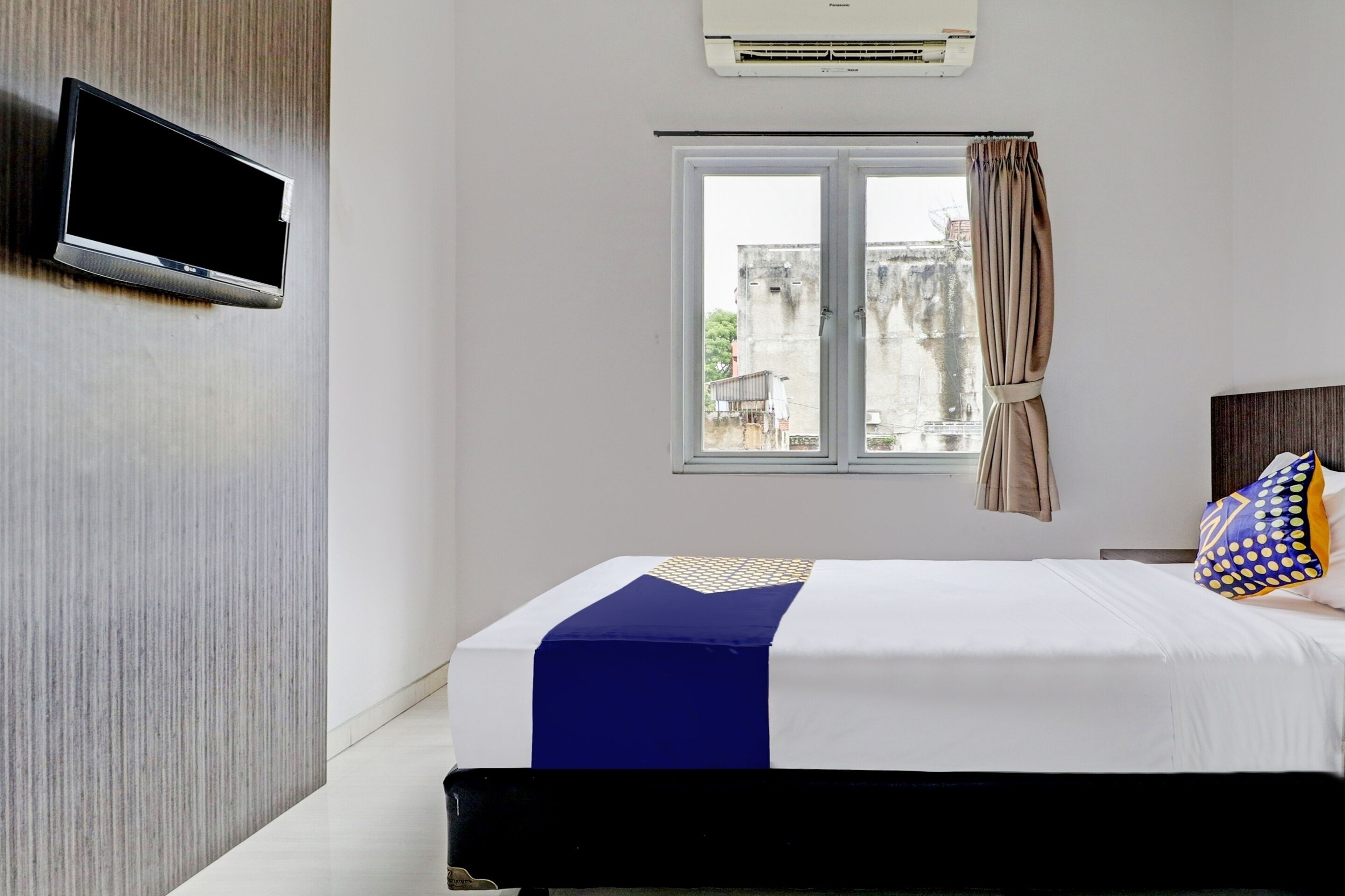 Een hotelkamer met een bed, een aan de muur bevestigde televisie, een raam met gordijnen en een airconditioning.