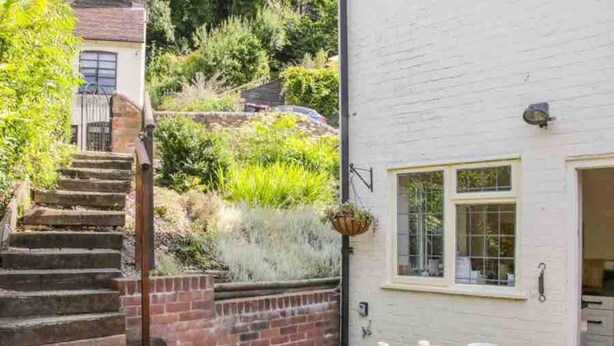 A white building with a small patio area featuring a round table with a teapot and cup, a brick staircase, and a hanging plant. - Cottagesleeps2-fireplace-valleyview-patio-garden (Telford)
