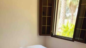 Un lit bien fait, avec du linge de lit et des serviettes blanches, une fenêtre donnant sur un paysage verdoyant et un sol carrelé.