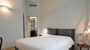 A neatly arranged bedroom with a large bed, a bedside table, a chair, and a desk with a lamp.