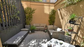 A small outdoor patio area with a sofa, a table set with teapots and cups, and potted plants.