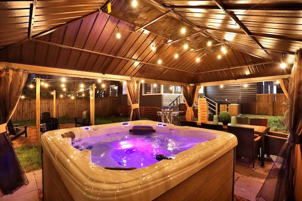 A hot tub with a wooden deck, string lights, and a covered area with outdoor furniture.
