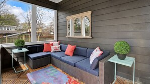 A modern outdoor seating area with a dark-colored sofa, two side tables with potted plants, and a large window offering a view of the neighborhood. - Modern Memphis Game House, Hot Tub! Close to Beale (Memphis)