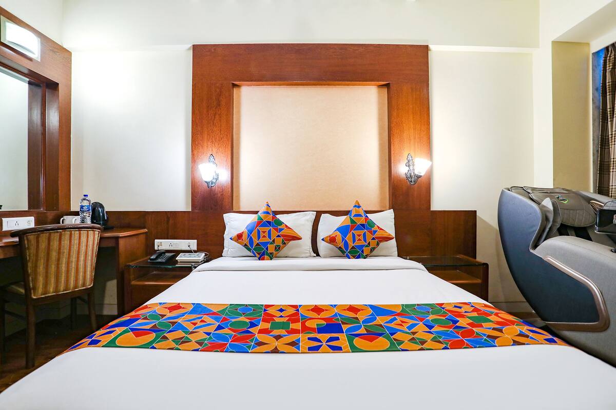 Habitación de hotel con cabecera de madera, dos almohadas decorativas, cama con colcha geométrica de colores, escritorio con silla y una mesita con teléfono y botella de agua.