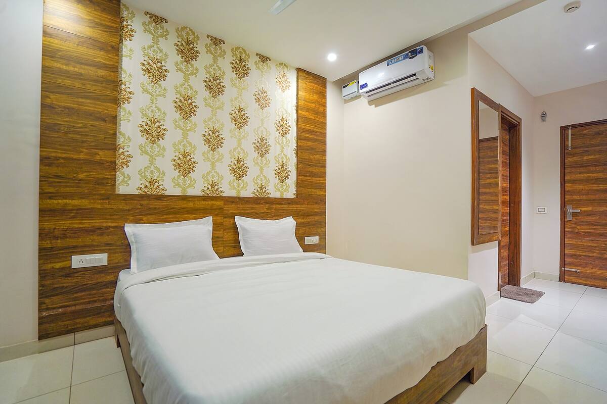 A hotel room with a wooden headboard, a large bed with white bedding, an air conditioning unit, and a wooden door.