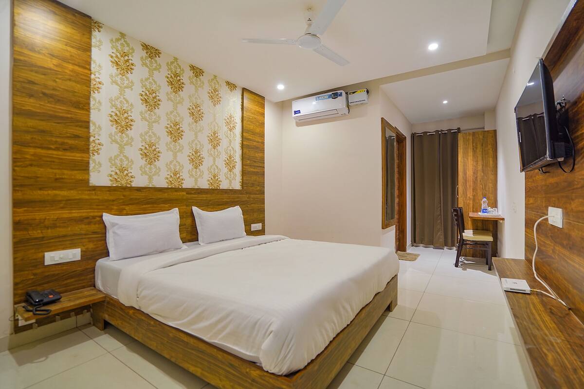 A hotel room with a wooden headboard, a bed with white linens, a ceiling fan, an air conditioning unit, a television mounted on the wall, and a small desk with a chair.