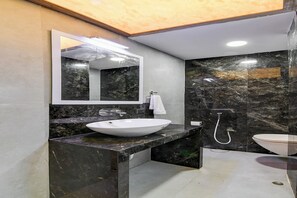 Un baño moderno con un espejo grande, un lavabo blanco, una ducha y paredes de mármol oscuro.
