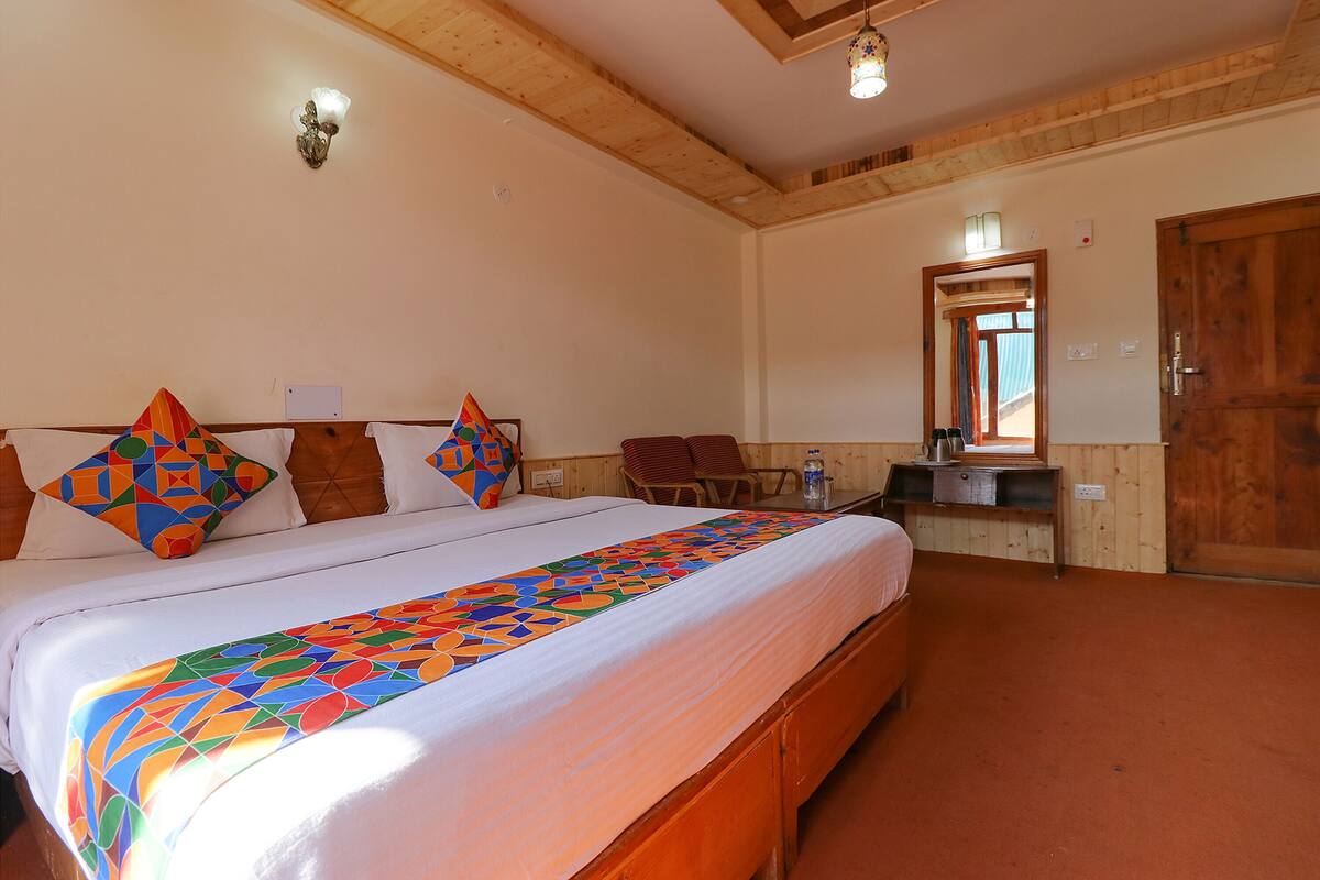 A bedroom with a large bed, wooden headboard, and a colorful geometric-patterned bedspread. There is a wooden dresser with a mirror and a small table with a lamp.