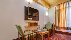 A cozy room with a wooden ceiling, a flat-screen TV mounted on the wall, two wooden chairs with cushions, a small wooden table, and a patterned carpet. - Fabhotel Shandela (Manali)