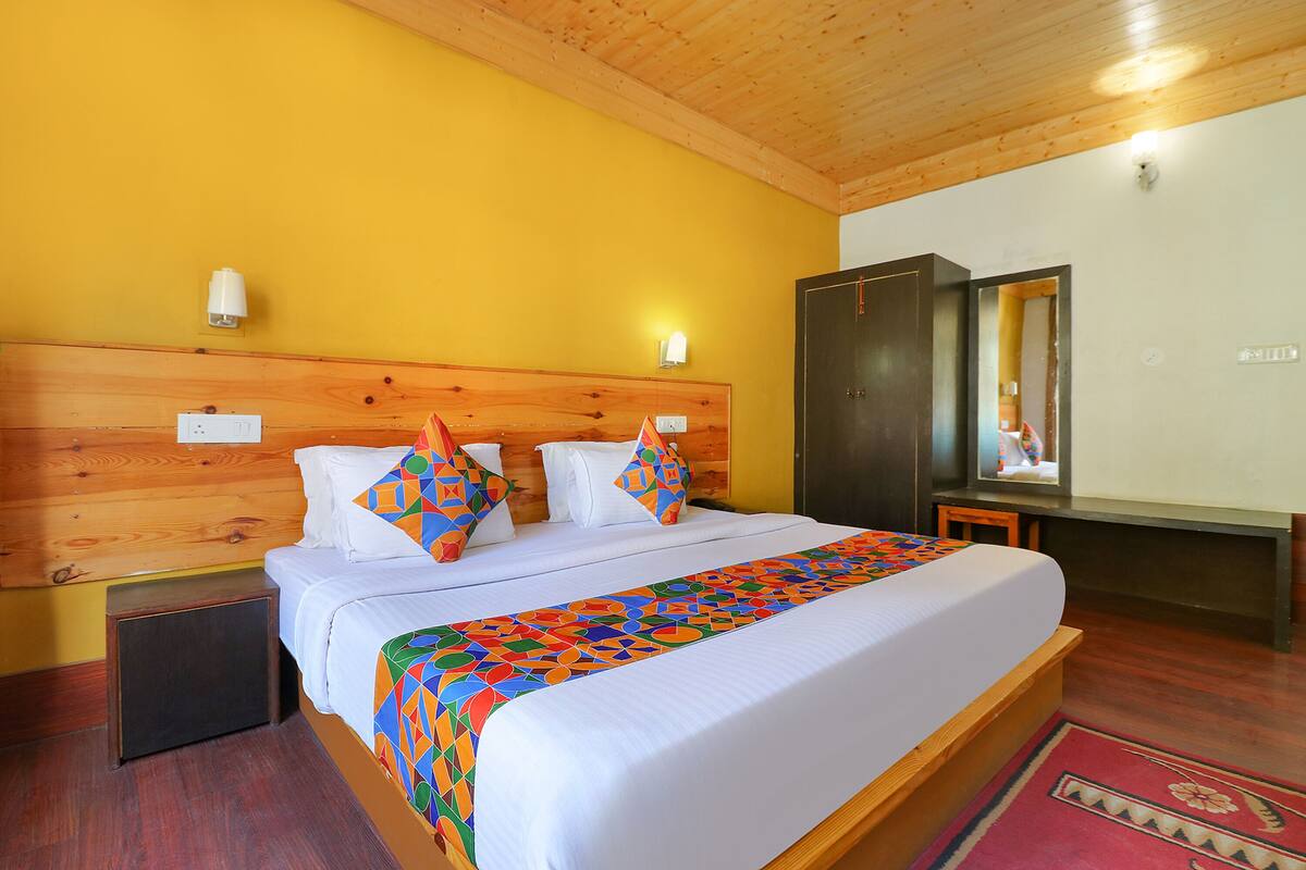 A hotel room with a wooden headboard, a bed with white bedding and colorful pillows, a small bedside table, and a wardrobe.