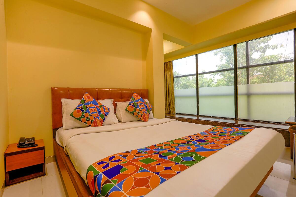 A hotel room with a bed, a nightstand, a window with curtains, and a view of greenery outside.