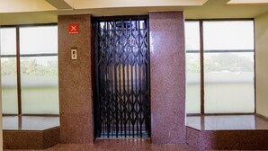 An elevator with a metal gate, a "No Smoking" sign, and large windows with a view of greenery and buildings. - Fabexpress Jayshree (Mumbai)
