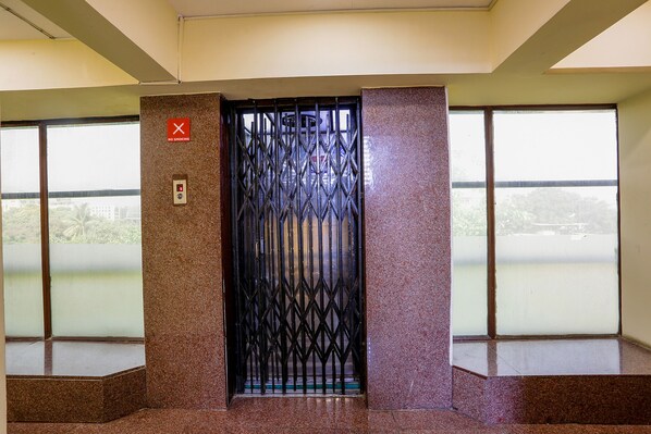An elevator with a metal gate, a "No Smoking" sign, and large windows with a view of greenery and buildings. - Fabexpress Jayshree (Mumbai)