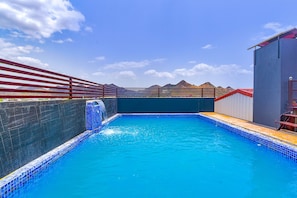 A swimming pool with a blue tiled edge, a red fence, and a mountain backdrop. - FabHotel Prime The Royal Galaxy (Udaipur)