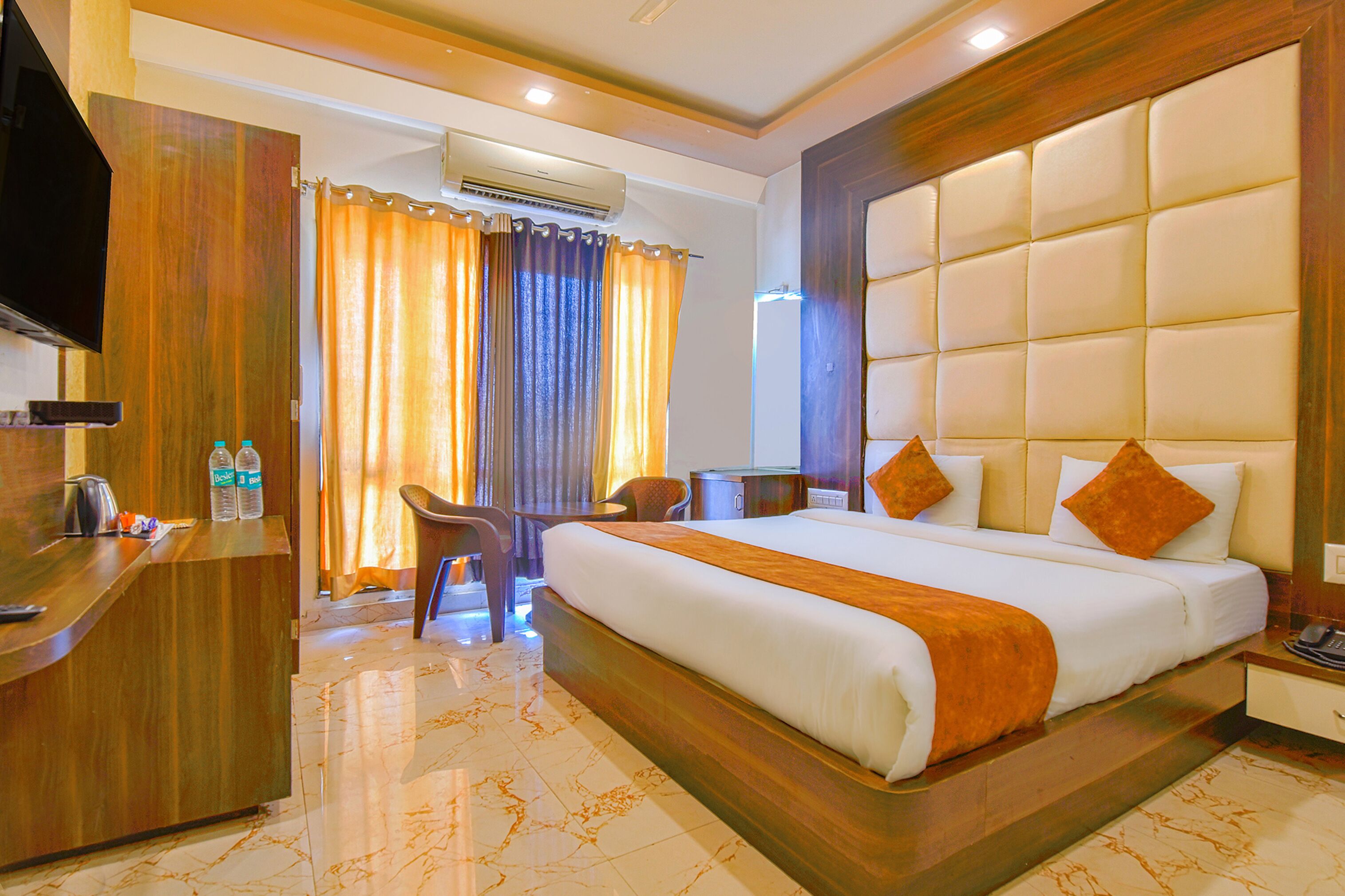 A hotel room with a large bed, a desk, a chair, an air conditioning unit, and a television.