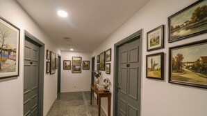 Un pasillo con pinturas de paisajes enmarcadas en las paredes, un banco de madera con un jarrón y libros, y una puerta con una placa de número.