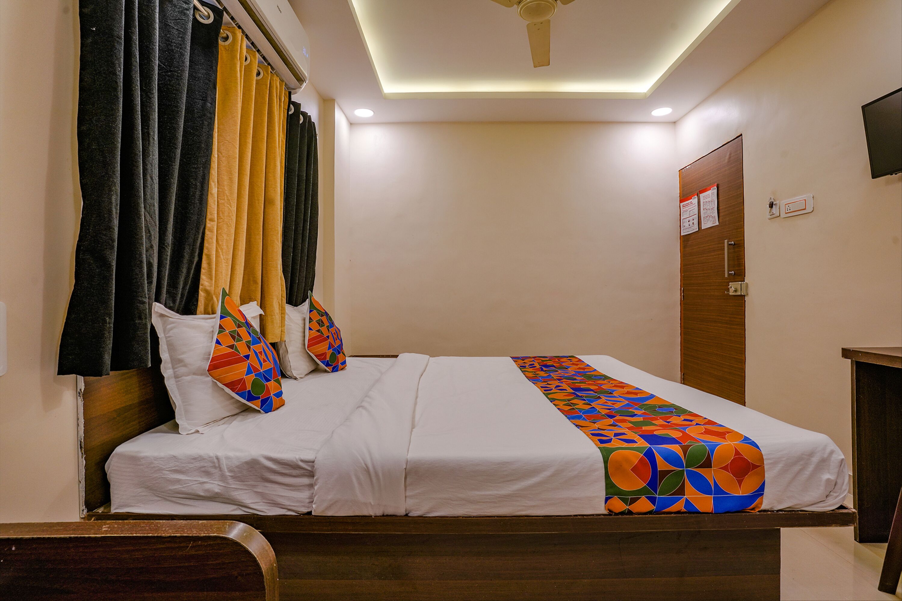 A hotel room with a bed, curtains, a ceiling fan, and a TV mounted on the wall.