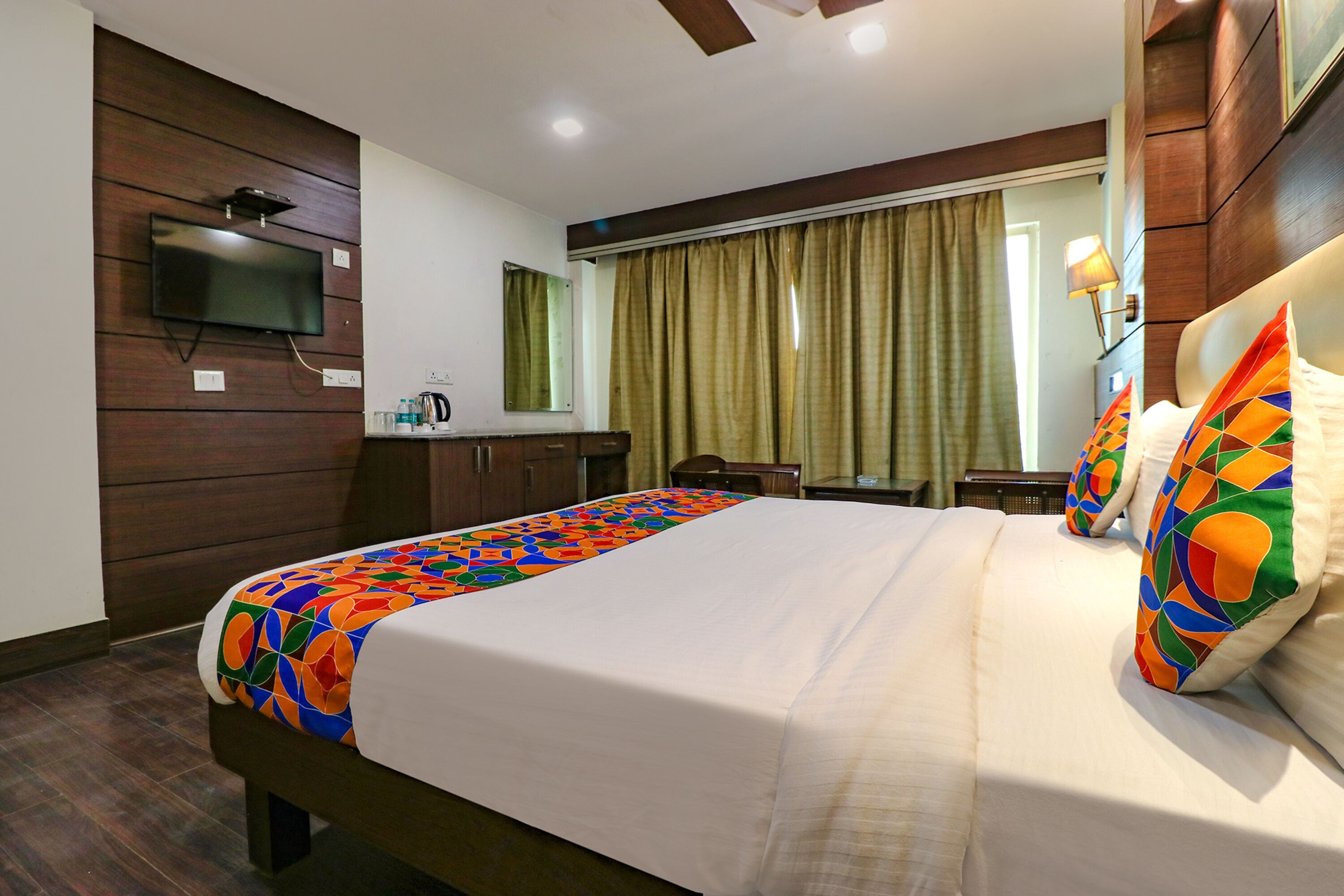 A hotel room with a bed, a TV mounted on the wall, a desk with a lamp, and a window with curtains.